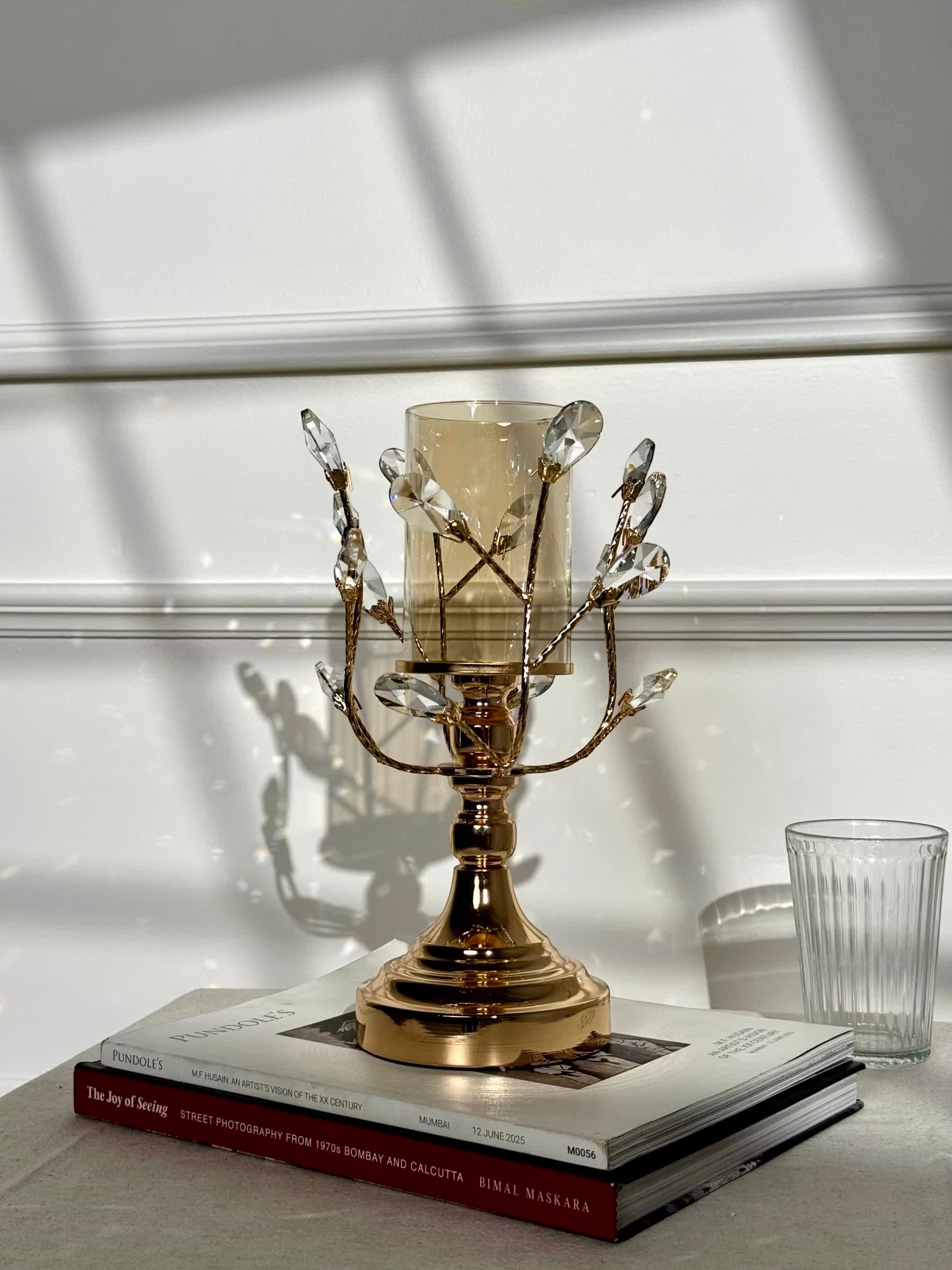 High Objects gold metal candle stand with crystal leaves and amber glass hurricane on books.