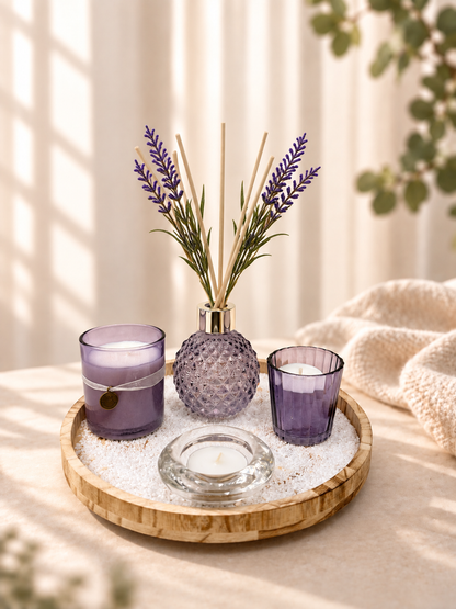 A styled home decor arrangement on a round wooden tray, featuring a purple textured glass reed diffuser bottle with lavender stems, two matching purple glass candle holders, a clear glass candle holder, and white decorative stones. The scene is set on a beige fabric surface with soft window shadows and a knitted blanket in the background.