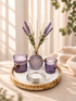 A styled home decor arrangement on a round wooden tray, featuring a purple textured glass reed diffuser bottle with lavender stems, two matching purple glass candle holders, a clear glass candle holder, and white decorative stones. The scene is set on a beige fabric surface with soft window shadows and a knitted blanket in the background.