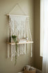 High Objects Anya Macramé Floating Pavilion shelf with ivory cotton cord, wood, and decorative fringe. Holds books and a plant.
