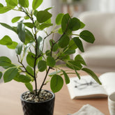 High Objects artificial plant with vibrant green leaves in a textured black pot, styled on a wooden table with a book and mug.