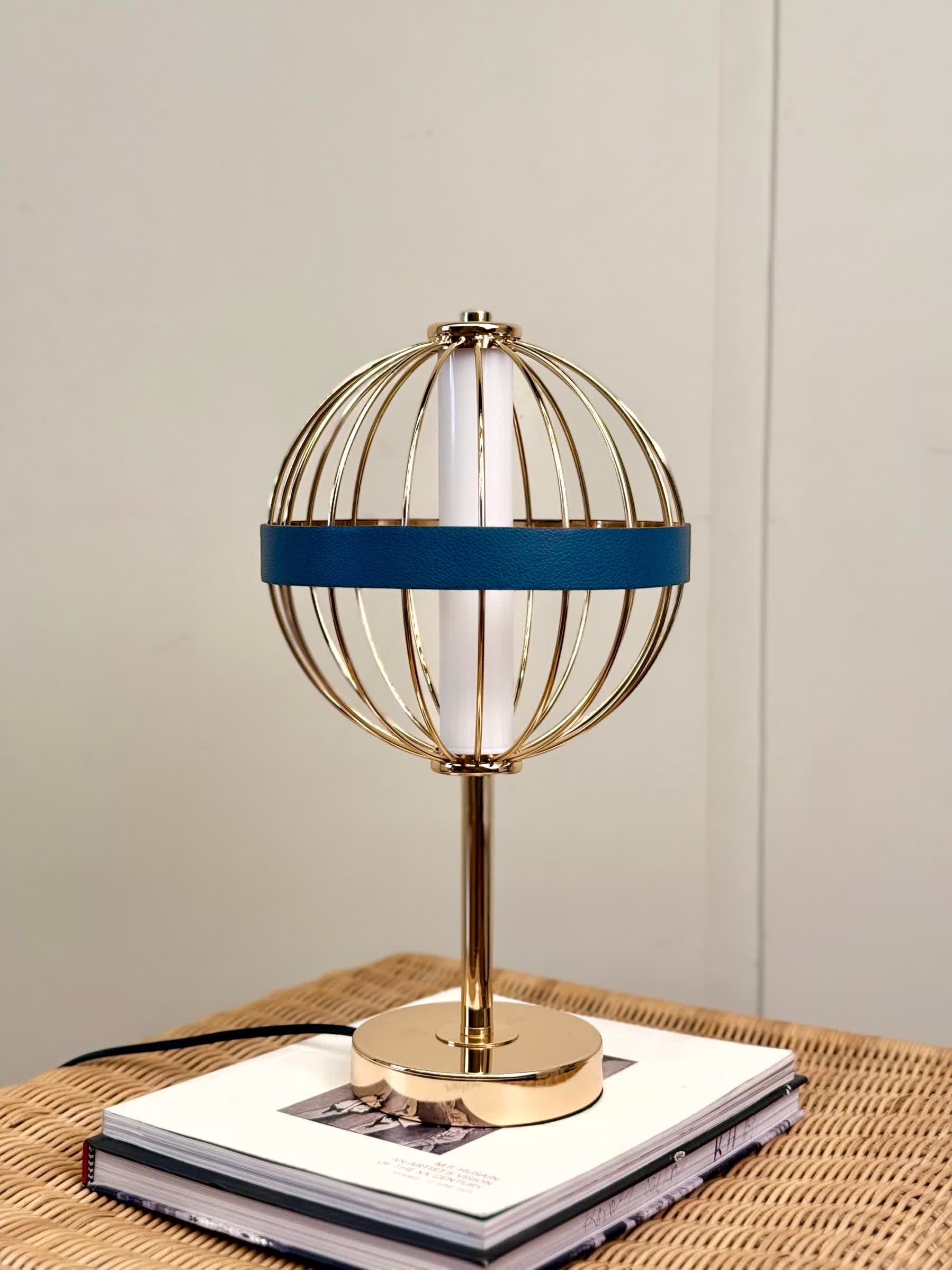 High Objects Azure Aurette Cage Table Lamp with gold wire globe, blue leather band, and gold stem on a stack of books.