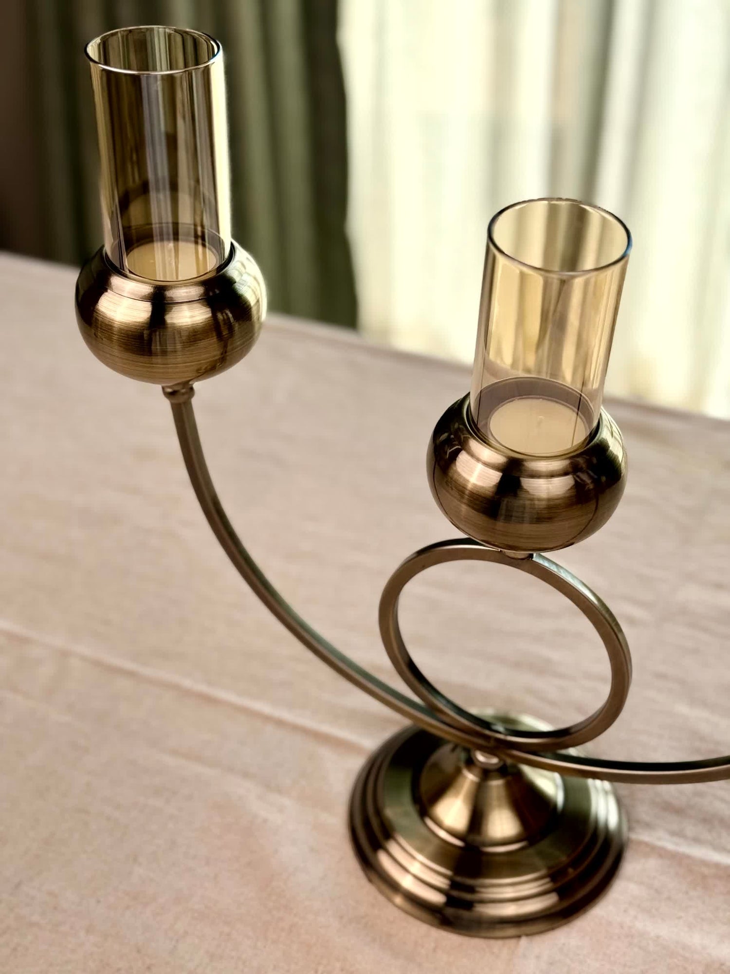 High Objects Circa Arc Triple Candle Stand in antique brass with amber glass shades. Features a sculptural arched frame and pedestal base.