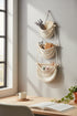 High Objects cream macrame wall organizer with 3 pockets, wood dowels, and hanging rope. Decorated with lavender, glasses, notebook, scissors, and spools.