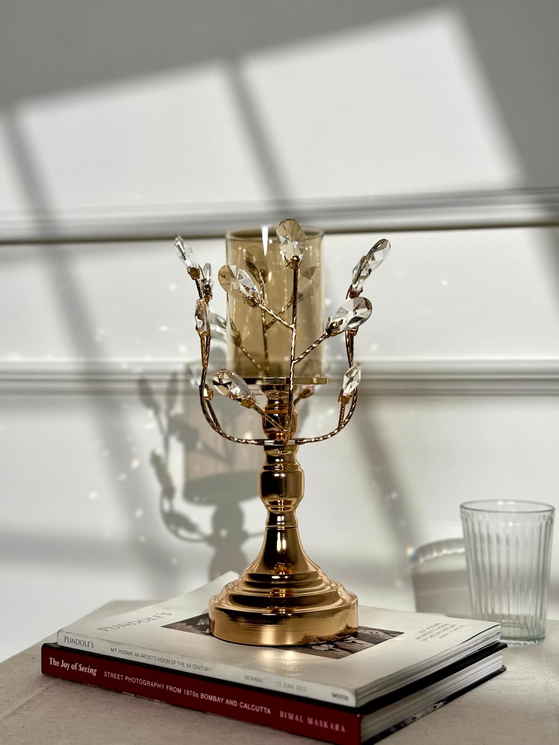 High Objects gold metal candle stand with crystal leaves and amber glass hurricane, on books.