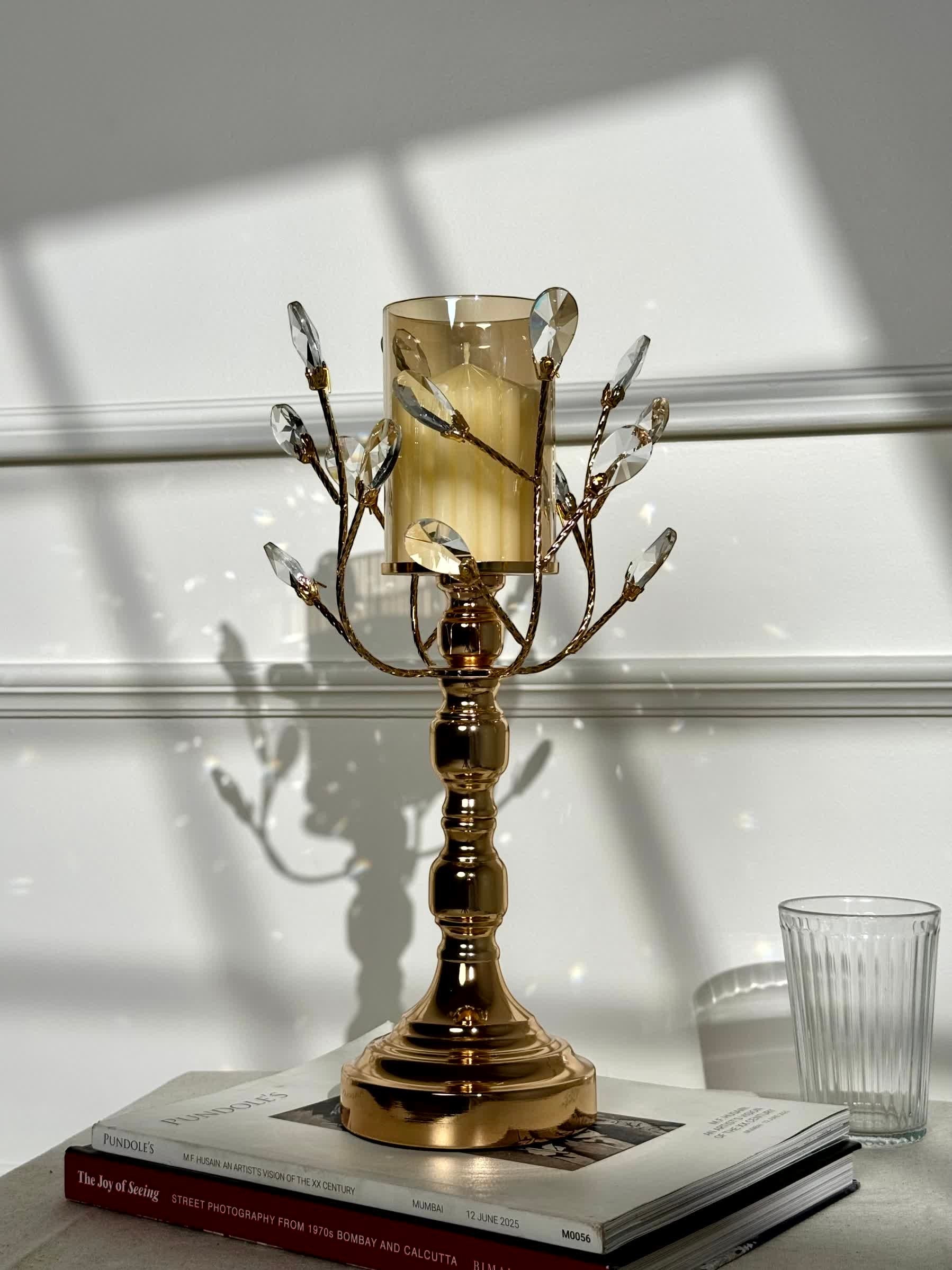 High Objects gold metal candle stand with crystal leaves and amber glass hurricane, holding a lit candle.