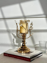 High Objects gold metal candle stand with crystal leaves and amber glass hurricane, holding a lit candle.