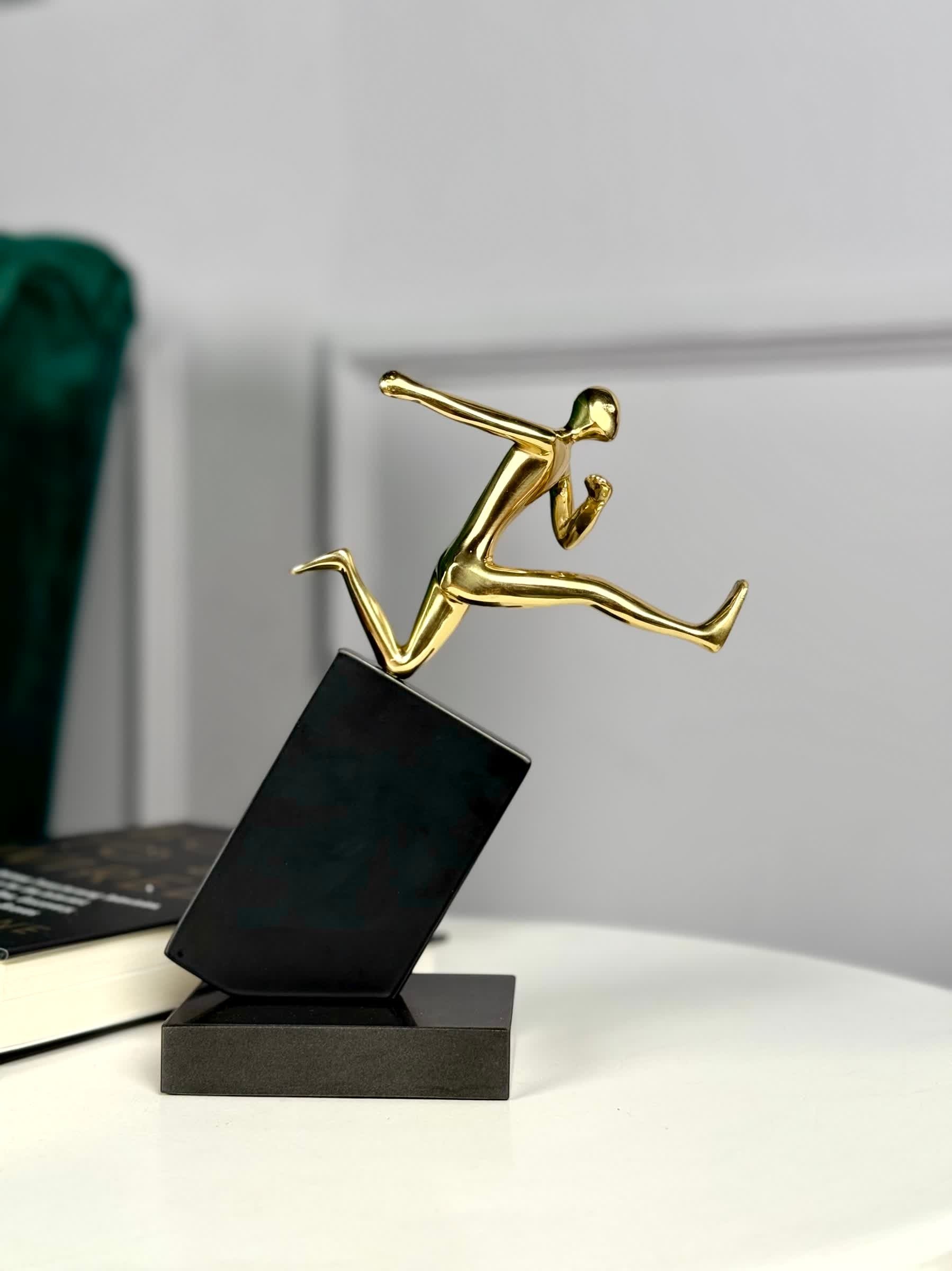 High Objects Golden Hurdler Leap Artefact sculpture, polished gold figure mid-flight on a black angled pedestal.