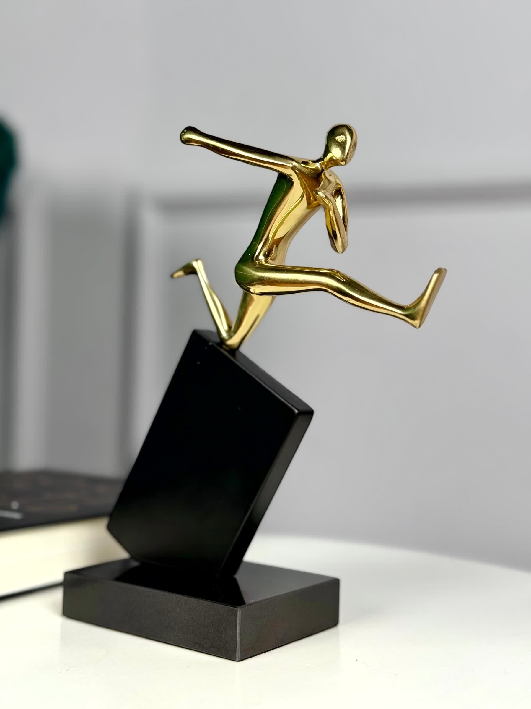 High Objects Golden Hurdler Leap Artefact sculpture, polished gold figure mid-flight on a black angled pedestal.