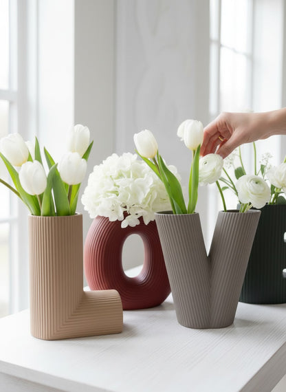 High Objects "L" vase in tan, "O" vase in burgundy, "V" vase in grey, and "E" vase in green, all with ribbed textures and holding white flowers.