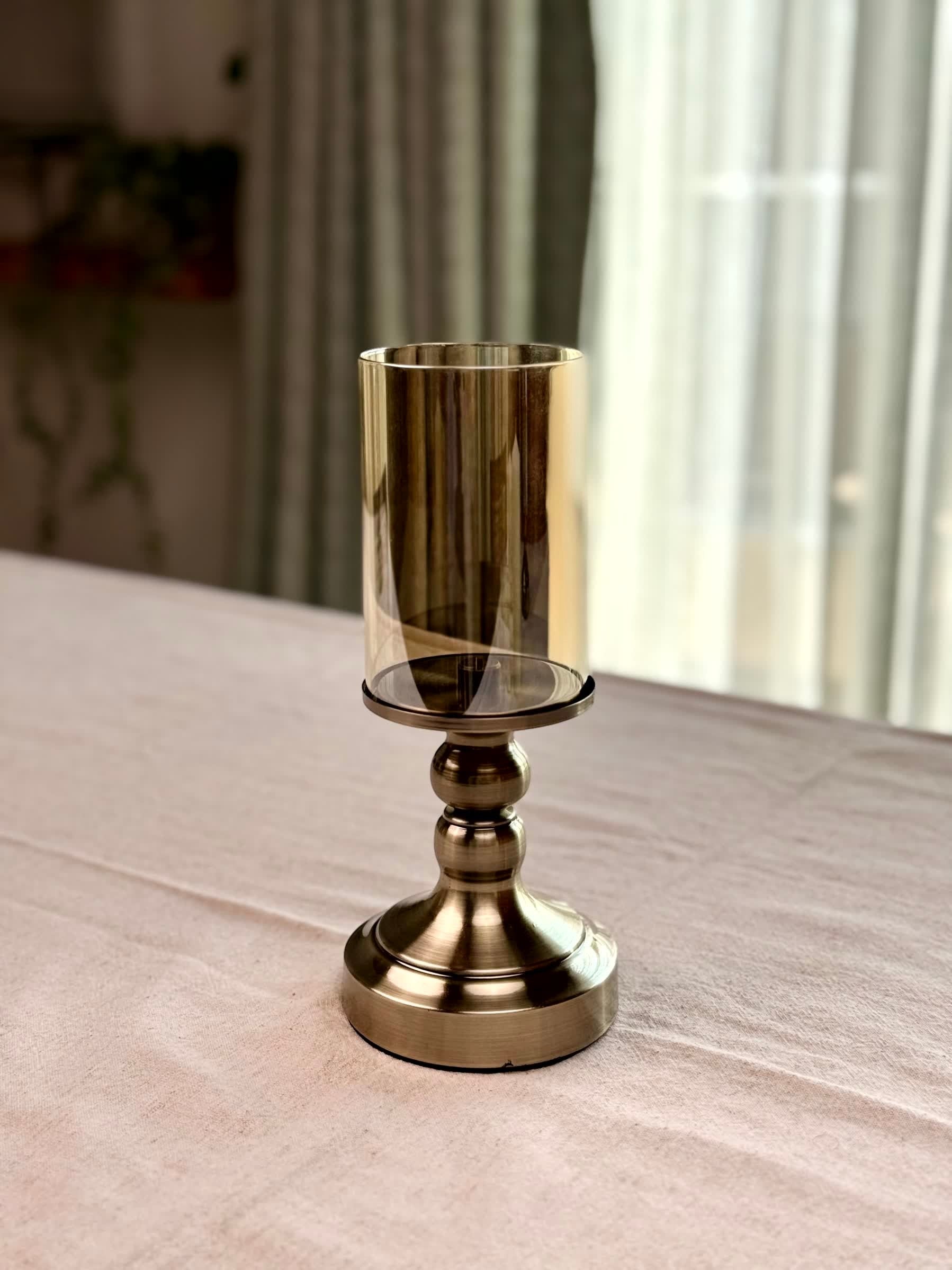 High Objects Luminaria Pedestal Candle Stand Set with tinted glass hurricane and metallic stem in antique brass finish.