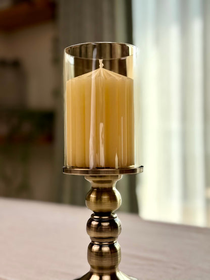 High Objects Luminaria Pedestal Candle Stand Set with a tall, fluted cream candle inside a clear glass hurricane and a bronze metallic stem.