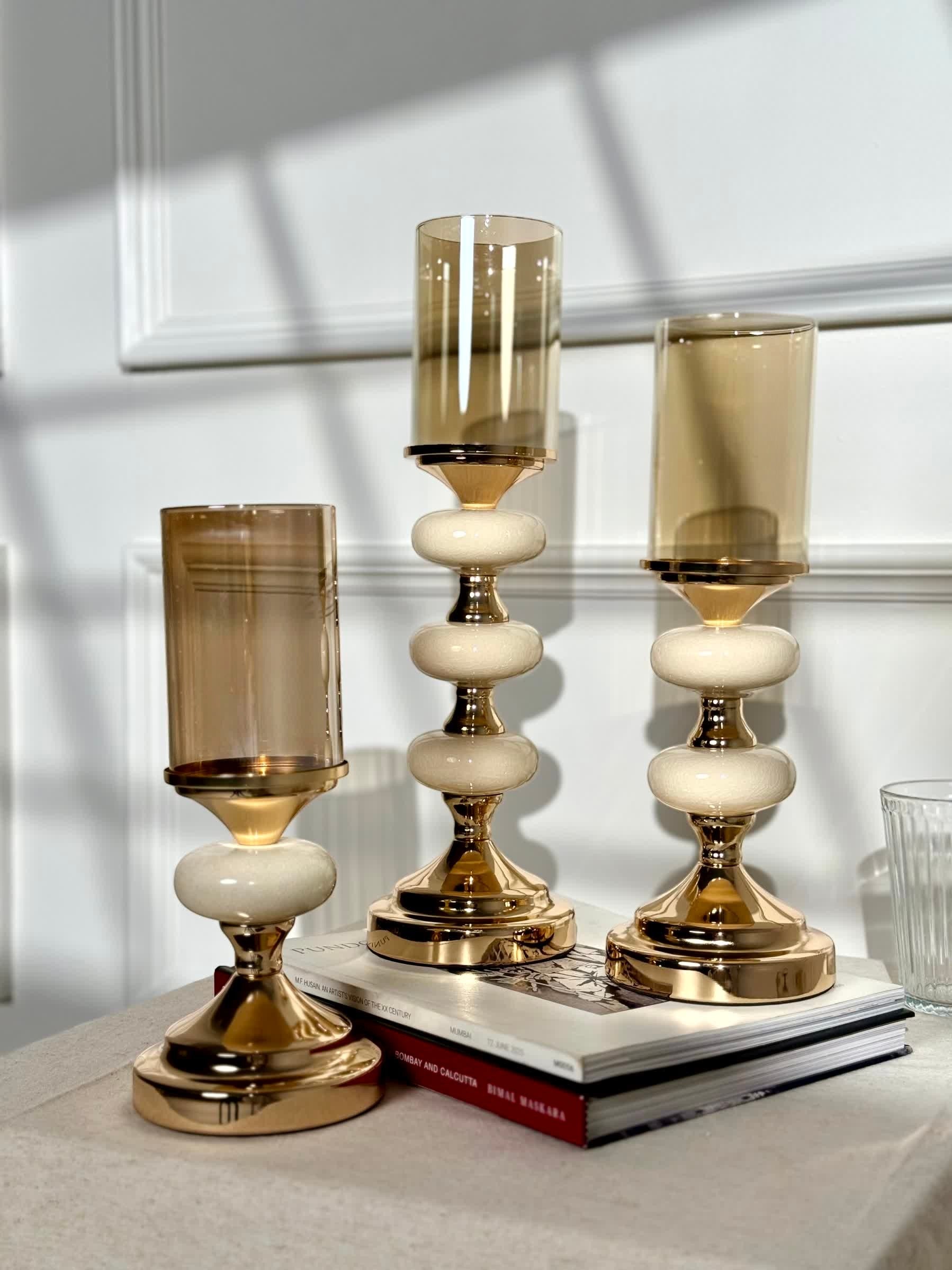 High Objects Pebble Luxe candle stand set with amber glass hurricanes and gold bases, featuring sculpted pebble-like stems.