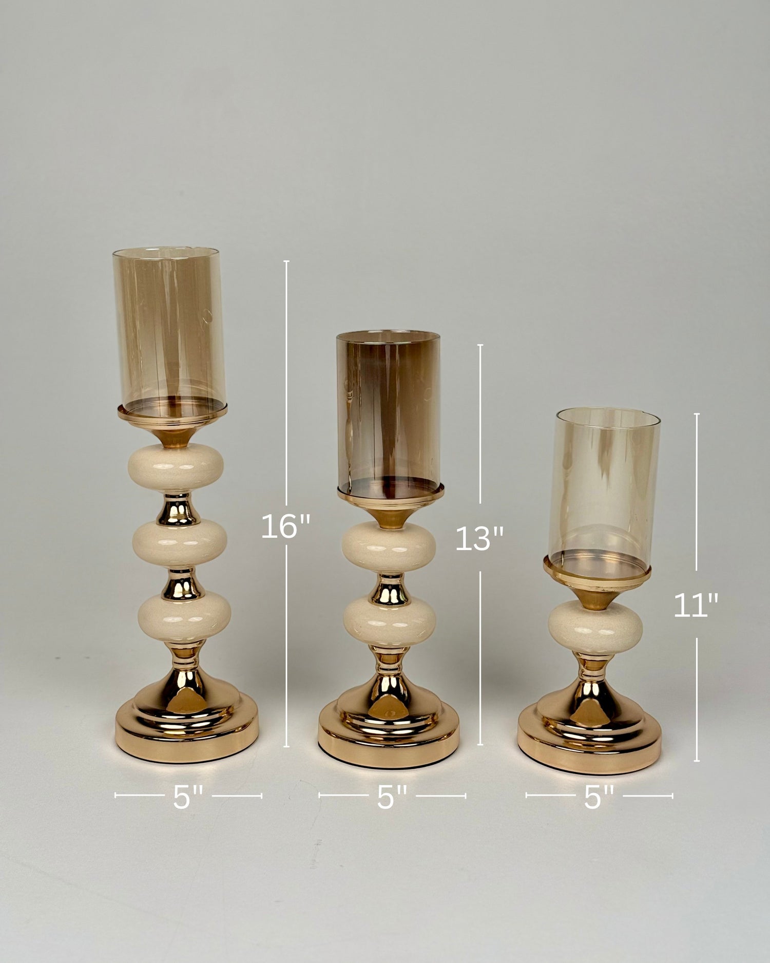 High Objects Pebble Luxe Candle Stand Set, set of 3 gold candle holders with amber glass hurricanes and cream pebble-like stems.