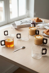 High Objects ORBITO™ set of 6 ribbed glass cups with dark geometric handles, two filled with latte art coffee, one with tea and lemon.