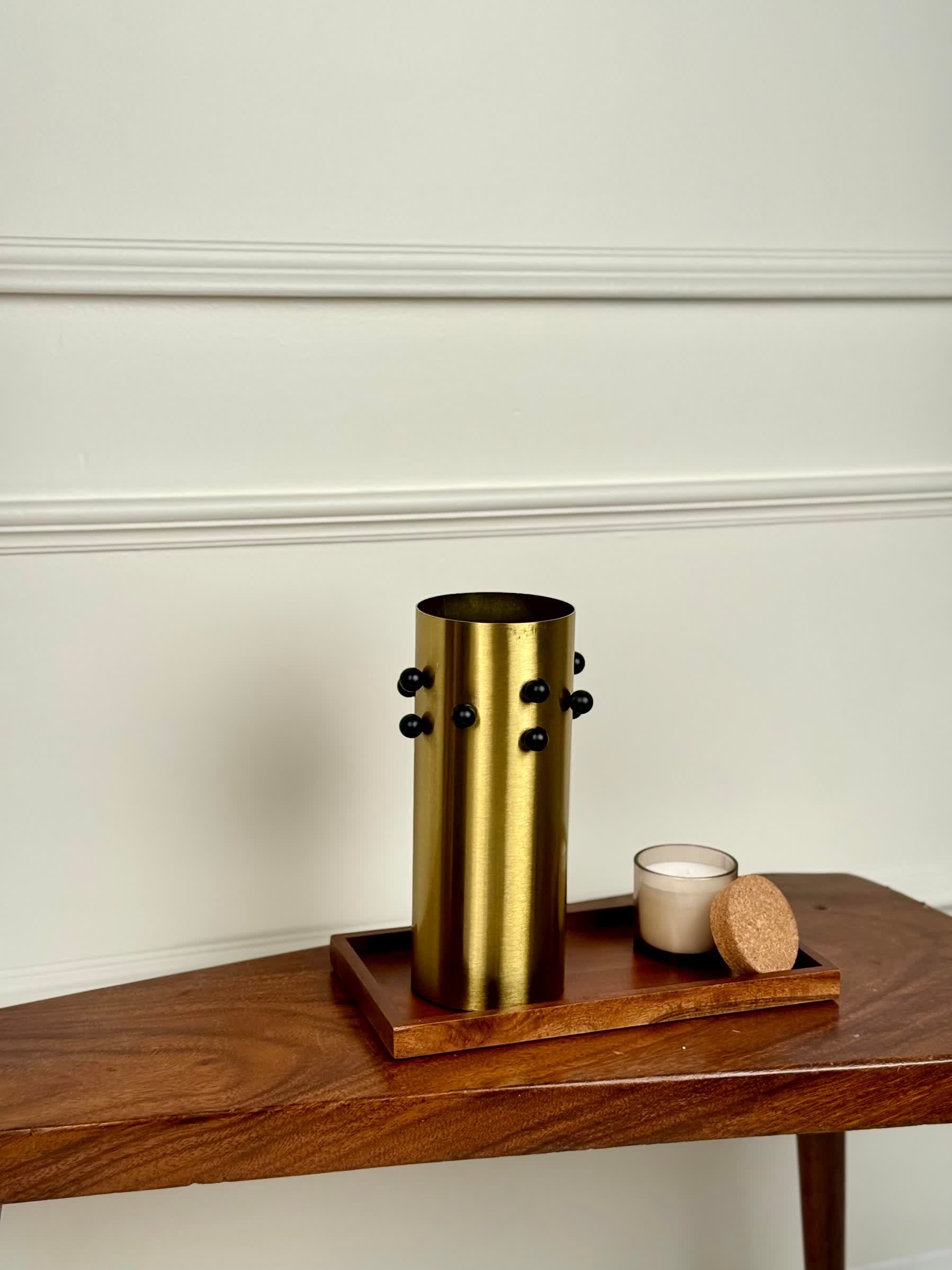 High Objects Studform Metal Vase, brushed gold cylindrical vase with black studs, on wooden tray with candle.