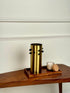 High Objects Studform Metal Vase, brushed gold cylindrical vase with black studs, on wooden tray with candle.