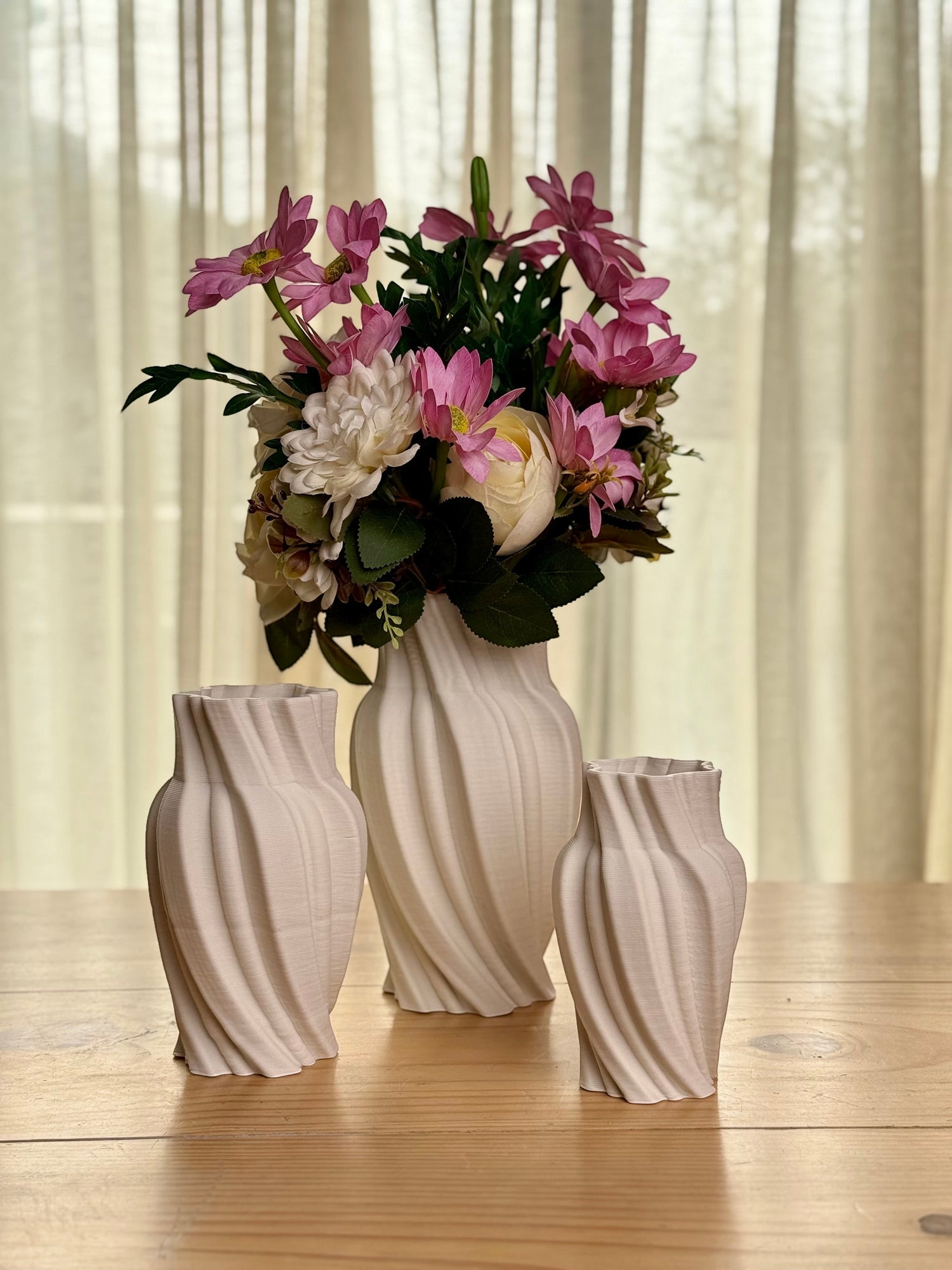 High Objects Volara Vase, a set of three 3D printed ceramic vases in matte white with a twisting, fluid design. The largest vase holds a bouquet of pink and white flowers.