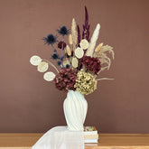 High Objects Volara Vase, a white ceramic vase with a twisted, fluid design, holding a bouquet of dried flowers in burgundy, blue, and cream.